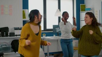 Authentic mixed raced happy women dancing cheerfully in startup creative agency office, during work hours, enjoying victory of construction project. Afterparty in workplace, colleagues having fun video