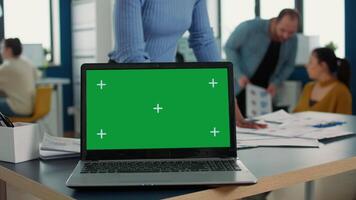 Laptop computer with green screen on desk in front of african american startup employee looking at paper with charts. Focus on portable computer screen with chroma key empty background. video