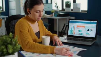 Portrait of startup employee analyzing financial data comparing paper with charts with statistics on laptop sitting at desk. Woman working in busy marketing department having morning coffee. video