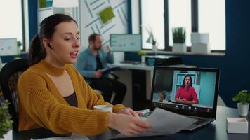 Portrait of startup employee presenting paper with charts using laptop in casual call conference with manager sitting at desk. Relaxed woman in busy department talking with executive. video