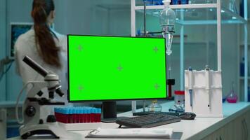 Desktop computer with green screen, mock up on display placed on desk in scientific laboratory while woman medical research scientist analysing virus evolution at digital monitor conducting experiment video
