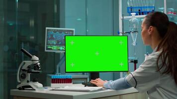 Over the shoulder shot of chemist working with green screen mock-up template on desktop computer, isolated display, chroma key. In background scietist doctor enter in medical lab with blood sample. video