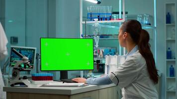 Female scientist working on computer with mock-up green screen in modern laboratory. Scientist doctor using various shelves with beakers, chemicals and different technical equipment is visible. video