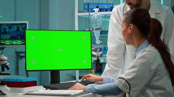 Medical research scientists using desktop computer with green screen mock up template in applied science laboratory. Lab engineers in white coats conduct experiments working with chroma key display video