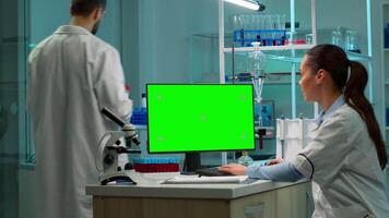 Biochemist sitting at workplace in laboratory using green mock-up screen personal computer with chroma key monitor while coworker working in background of pharmaceutical research centre. video