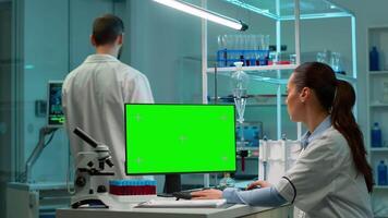 forskare Sammanträde på skrivbord arbetssätt på personlig dator med mock-up grön skärm. i bakgrund man labb forskare diskuterar med läkare handla om vaccin utvecklande föra blod prover video