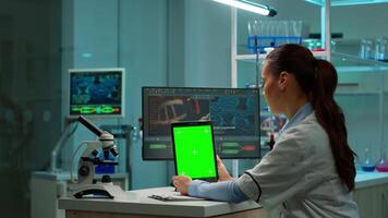 kvinna kemist forskare använder sig av grön mock-up skärm läsplatta Sammanträde på skrivbord. i bakgrund teknologi forskning, utveckling laboratorium med specialist läkare arbetssätt i hög tech design video