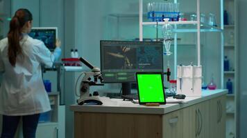Microbiologist working on monitor in background analysing test samples with tablet with green chroma key screen placed on desk in front. Team of biotechnology scientists developing drugs. video