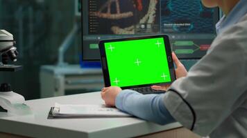 tillbaka se av kemist kvinna innehav läsplatta med grön attrapp i modern utrustad labb. team av mikrobiologer håller på med vaccin forskning skrivning på enhet med krom nyckel, isolerat visa. video
