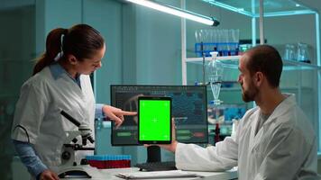 Medical research scientists using notepad with green screen mock up template in applied science laboratory pointing on chroma key display.. Lab engineers in white coats conduct experiments working video