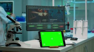 Display tablet with green screen, mock up on template placed on desk in scientific laboratory while woman medical research scientist analysing virus evolution at digital monitor conducting experiment video