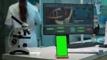 Display phone with green screen, mock up on template placed on desk in scientific lab while team of medical researcher scientists analysing virus evolution at digital monitor conducting experiment video