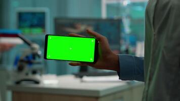 Microbiologist holding smartphone with green chroma key display in front of camera in modern equipped lab. Team of biotechnology scientists developing drugs using tablet with mock up screen. video