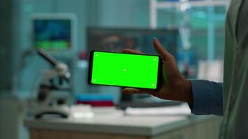 stänga upp av forskare kvinna innehav horisontell telefon med grön attrapp i modern utrustad labb. team av mikrobiologer håller på med vaccin forskning skrivning på enhet med krom nyckel, isolerat visa. video