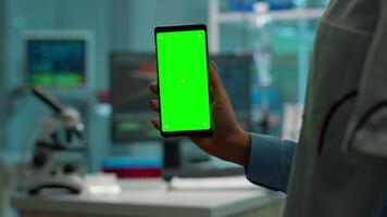 krom nyckel isolerat visa på smartphone Begagnade förbi forskare kvinna i labb skåp och kollegor i vit täcka föra blod prov. forskare använder sig av mobil telefon med attrapp grön skärm video