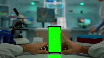 pOV skott av kemist använder sig av smartphone med grön skärm i biologisk laboratorium. medicinsk arbetstagare bär vit täcka i klinik arbetssätt med mobil med krom nyckel på isolerat visa i medicinsk labb video