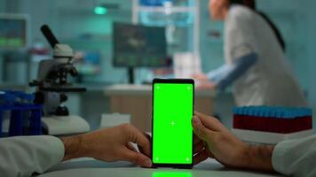 pOV skott av man forskare Sammanträde på skrivbord arbetssätt på mobil telefon med mock-up grön skärm, isolerat visa. i bakgrund labb forskare analyserar vaccin utvecklande granskning prover video