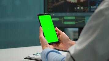 Medic in scientific laboratory holding phone with greenscreen wearing white coat while nurse bringing blood samples. Healthcare specialist in modern lab using smartphone with mockup chroma key display video