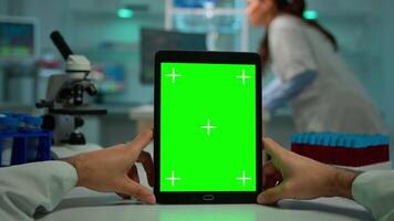 Pov shot of man scientist sitting at desk working on notebook with mock-up green screen, isolated display. In background lab researcher analysing vaccine developent examining samples video
