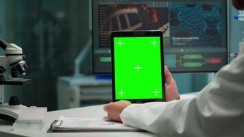 Close up of biochemist sitting at workplace in laboratory using green mock-up screen tablet with chroma key display while coworker working in background bringing blood sample. video