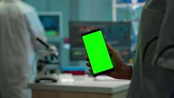 stänga upp av biokemist innehav och ser på telefon med krom nyckel visa stående i biologisk labb medan sjuksköterska i vit täcka föra blod prov. forskare använder sig av telefon med mockup, grön skärm video