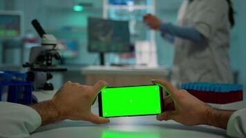 pOV skott av kemist använder sig av horisontell smartphone med krom nyckel på isolerat visa i biologisk laboratorium. läkare bär vit täcka i klinik arbetssätt med mobil med grön skärm i medicinsk labb video