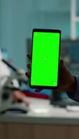 Vertical . Close up of chemist's hand holding smartphone with green screen standing in biological laboratory while nurse bringing blood samples. Scientist using smartphone with mockup, chroma key display video