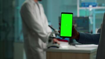 stänga upp av apotek hand innehav smartphone med grön skärm stående i biologisk laboratorium medan sjuksköterska föra blod prover. forskare använder sig av smartphone med mockup, krom nyckel visa video