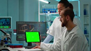 Man research scientist has conversation with famale bioengineer next to a desktop computer holding tablet with green screen mock up. They look at chroma key pc template in a modern science laboratory. video