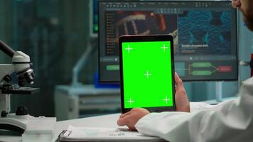 Male chemist scientist holding green mock-up screen tablet sitting at desk. In background technology research, development laboratory with specialist doctor working in high tech design video