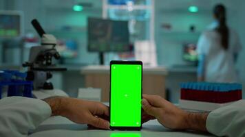 pOV skott av mikrobiolog innehav telefon med grön krom nyckel visa Sammanträde på skrivbord sökande, läsning virus symtom. i bakgrund labb forskare analyserar vaccin utvecklande granskning prover video