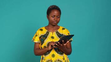 Natural cute girl browsing internet on a tablet against blue background, navigating on online web pages by using a portable device. Woman posing with a gadget and traditional outfit. Camera A. video
