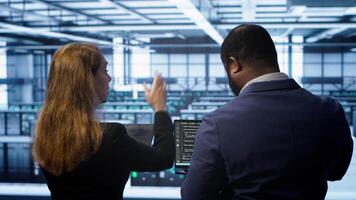 Teamworking programmers in data center overseeing server operations using laptop and tablet. Coworkers ensuring optimal performance and data integrity in server room using devices, camera A video