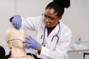 A female medical professional wearing a white coat and stethoscope practices applying an oxygen mask to a mannequin photo