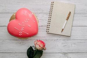 Heart shaped pink mousse cake on table with rose and notepad. photo