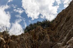 View from below of a rocky cliff with vegetation above against a blue sky and white clouds. Copy space.nature or abstract content, photo
