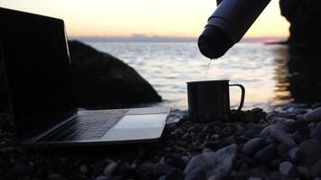 Silhouette of laptop and tea is poured into a cup on the beach over sea sunset. Sparkling sea water on background. Freelancer, blogger working on notebook on the beach. video