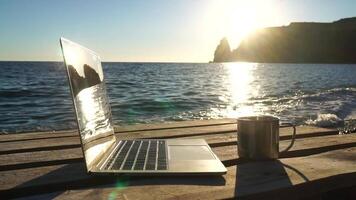 Silhouette of laptop and cup on the beach over sea sunset. Sparkling sea water on background. Freelancer, blogger working on notebook on the beach. video