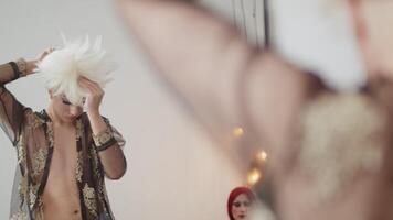 Young man in drag queen makeup putting on a wig on his head in front of the mirror and adjusting it in the dressing room during preparation for the performance video