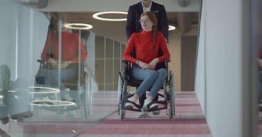 A company director assists his business colleague in a wheelchair, helping her navigate to their startup office, where they work alongside their diverse team of colleagues, emphasizing inclusivity and video