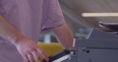 A modern, blond-haired man efficiently operates a high-tech paper printer in a large, contemporary office, showcasing advanced office technology and workflow management video