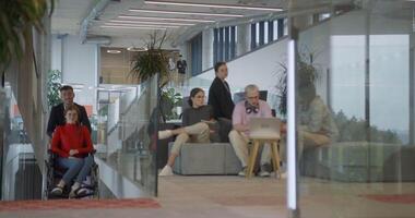 A company director assists his business colleague in a wheelchair, helping her navigate to their startup office, where they work alongside their diverse team of colleagues, emphasizing inclusivity and video
