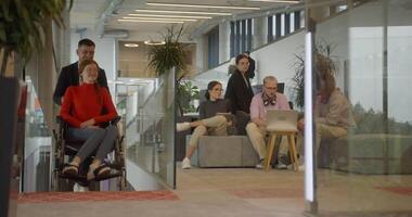 A company director assists his business colleague in a wheelchair, helping her navigate to their startup office, where they work alongside their diverse team of colleagues, emphasizing inclusivity and video