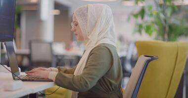 A businesswoman in a hijab is seen working on a computer in a modern office, highlighting inclusivity and professionalism in a diverse workplace video