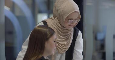 A diverse group of businesswomen, one of whom wears a hijab, collaborates while focusing on a laptop, engaging in discussions and problem-solving, showcasing professionalism, teamwork, and effective video