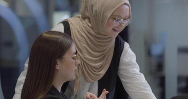 A diverse group of businesswomen, one of whom wears a hijab, collaborates while focusing on a laptop, engaging in discussions and problem-solving, showcasing professionalism, teamwork, and effective video