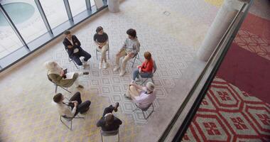 Top view of a diverse group of young business entrepreneurs gathered in a circle for a meeting, discussing corporate challenges and innovative solutions within the modern confines of a large video