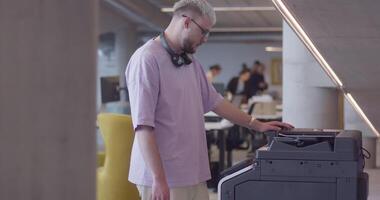 A modern, blond-haired man efficiently operates a high-tech paper printer in a large, contemporary office, showcasing advanced office technology and workflow management video