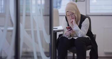 A businesswoman in a hijab takes a break from her work in a modern office, using her smartphone in a contemporary workspace, highlighting diversity, empowerment, and the balance of technology and video