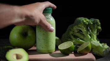 Refreshing green smoothie in bottles on wooden board. Hand picks up smoothie from table with avocado, lime, broccoli, apple, cucumber. Perfect for healthy lifestyle content. Ideal smoothie video